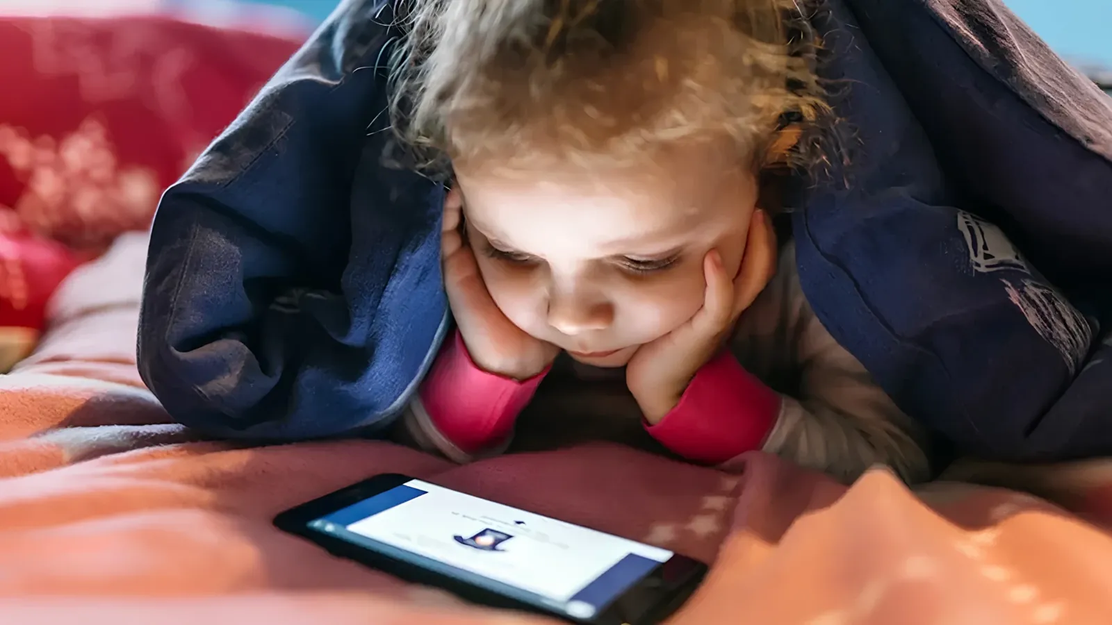 Dieťa leží v posteli a pozerá do mobilu pod perinou